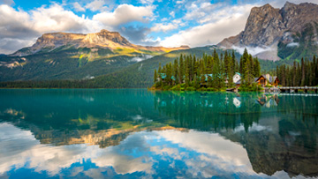 Un lac de montagne serein aux eaux turquoise cristallines, dans lesquelles se reflètent les pins environnants, les chalets et les sommets escarpés, sous un ciel partiellement nuageux, où se mêlent des verts éclatants, des bleus et des tons terreux, avec un petit ponton s'avançant dans l'eau.